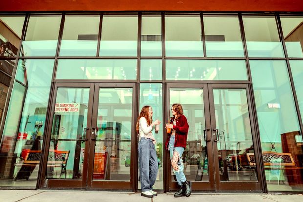 Two people stand talking outside a building with glass doors, holding drinks.