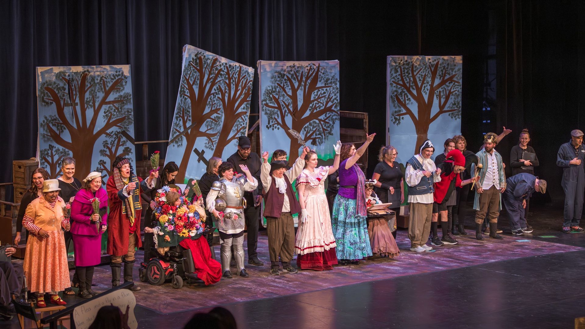 Theater cast bows on stage, backdrop of trees. Bright costumes, diverse group, audience seating visible.