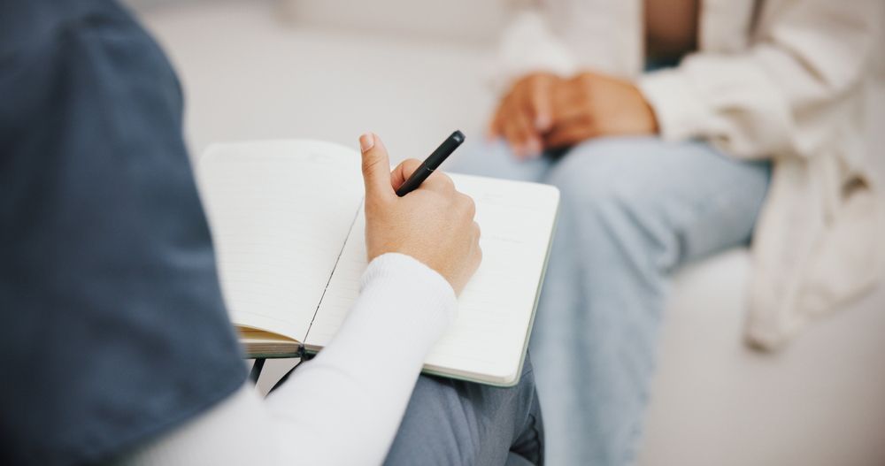 A person writing in a notebook, interviewing another person sitting nearby.
