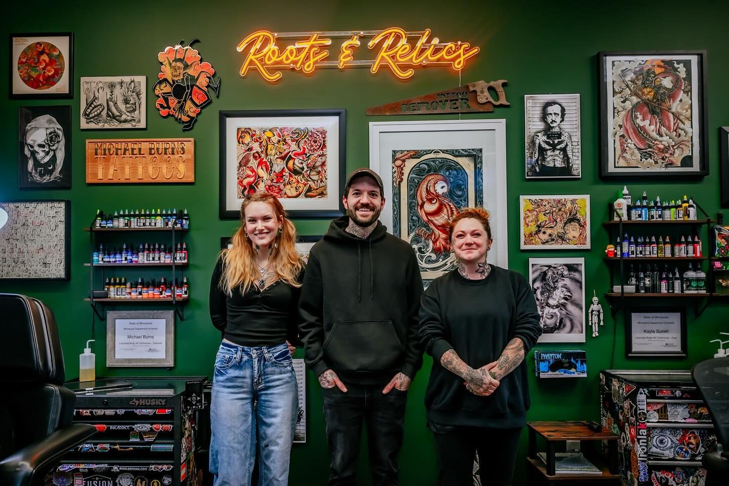 Three people stand in a tattoo parlor with green walls and art. A neon sign reads 