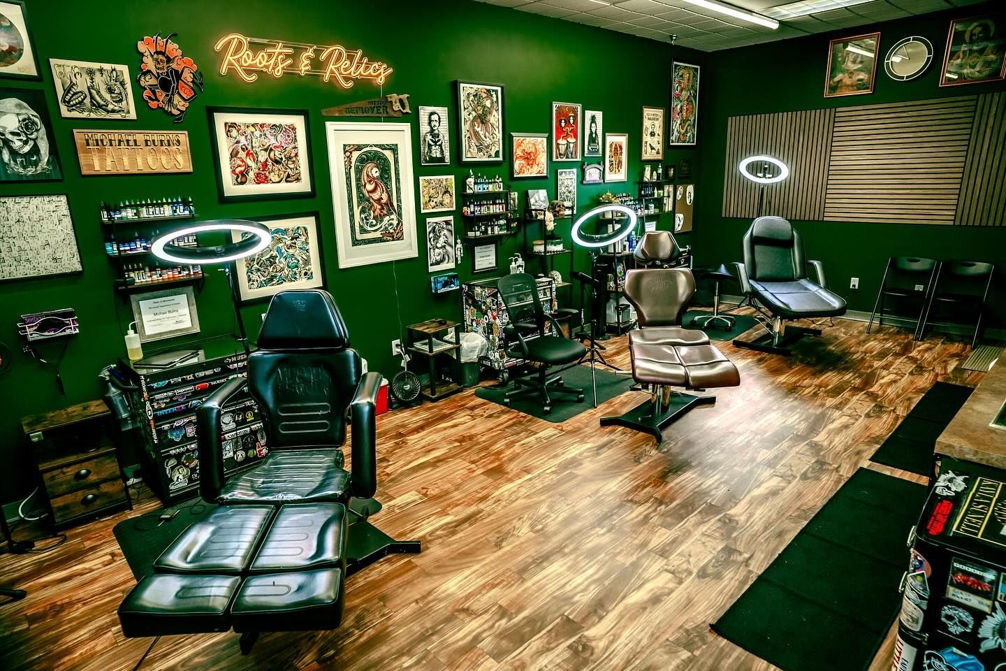 Tattoo studio interior with green walls, wooden floor, tattoo chairs, artwork, and bright ring lights.