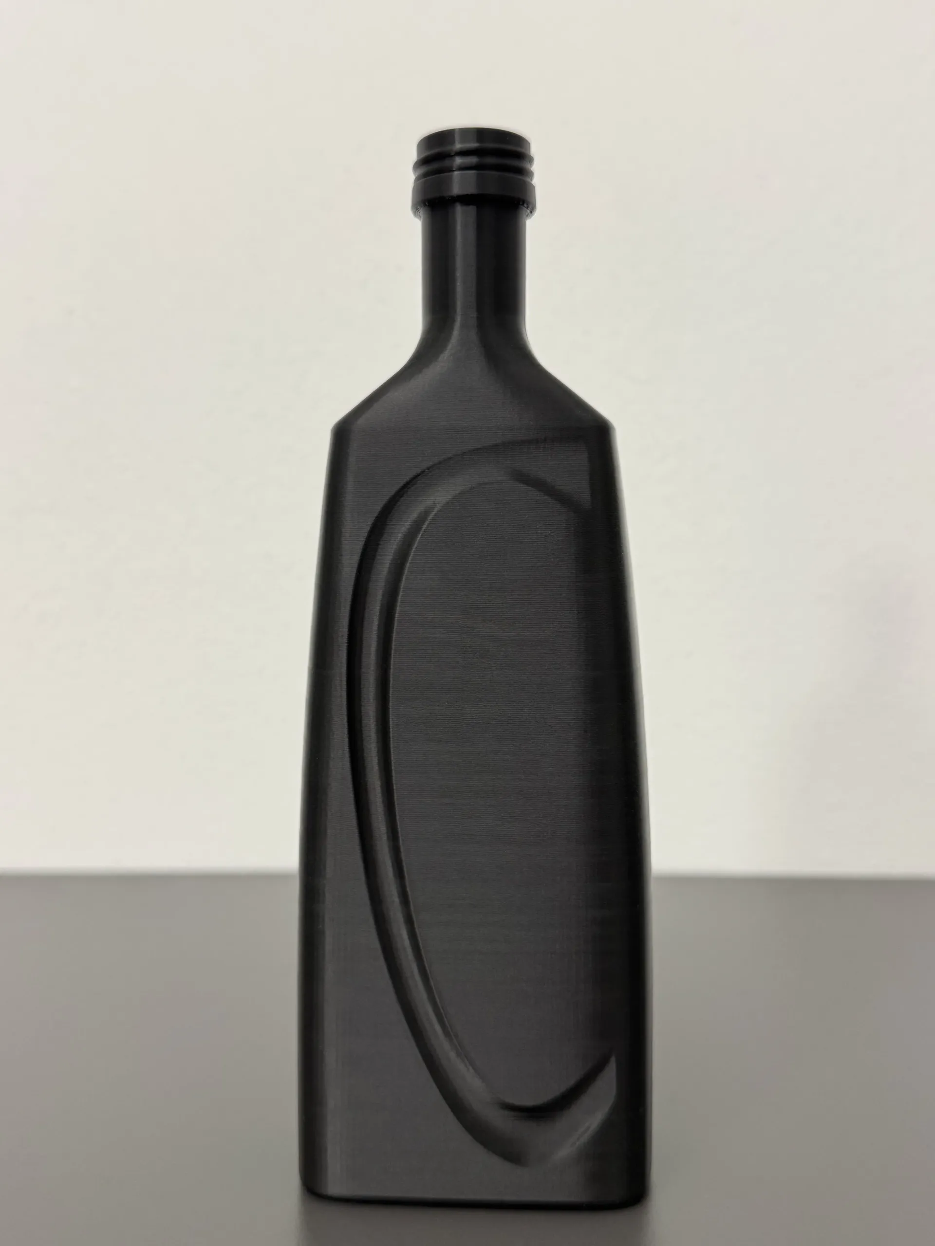 Bottiglia rettangolare nera e testurizzata con una scanalatura curva sulla parte anteriore, su uno sfondo grigio semplice.