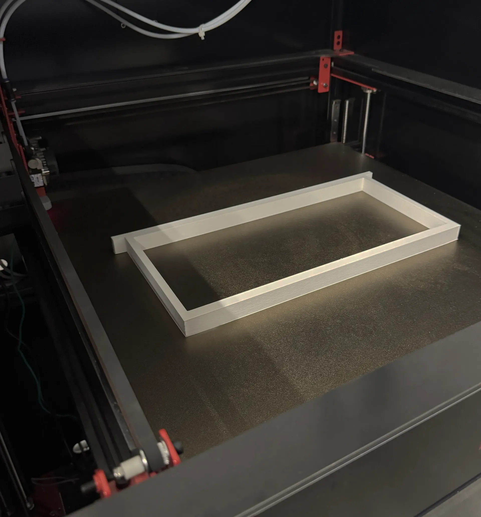 Una stampante 3D in funzione, che costruisce una cornice rettangolare con materiale bianco su un letto riscaldato.