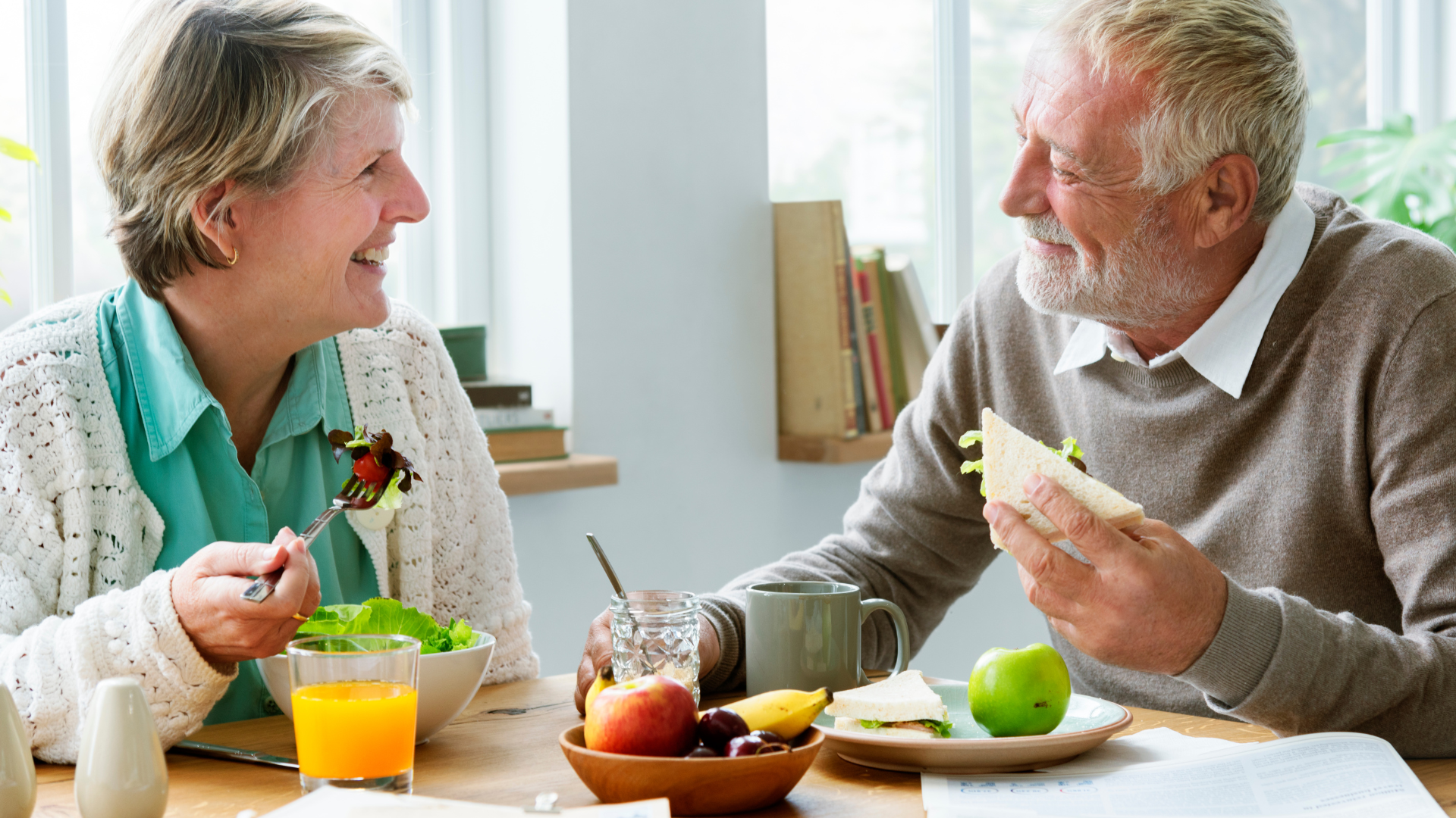 Older couple smiling and eating at a table by a window; she has salad, he has a sandwich and fruit.