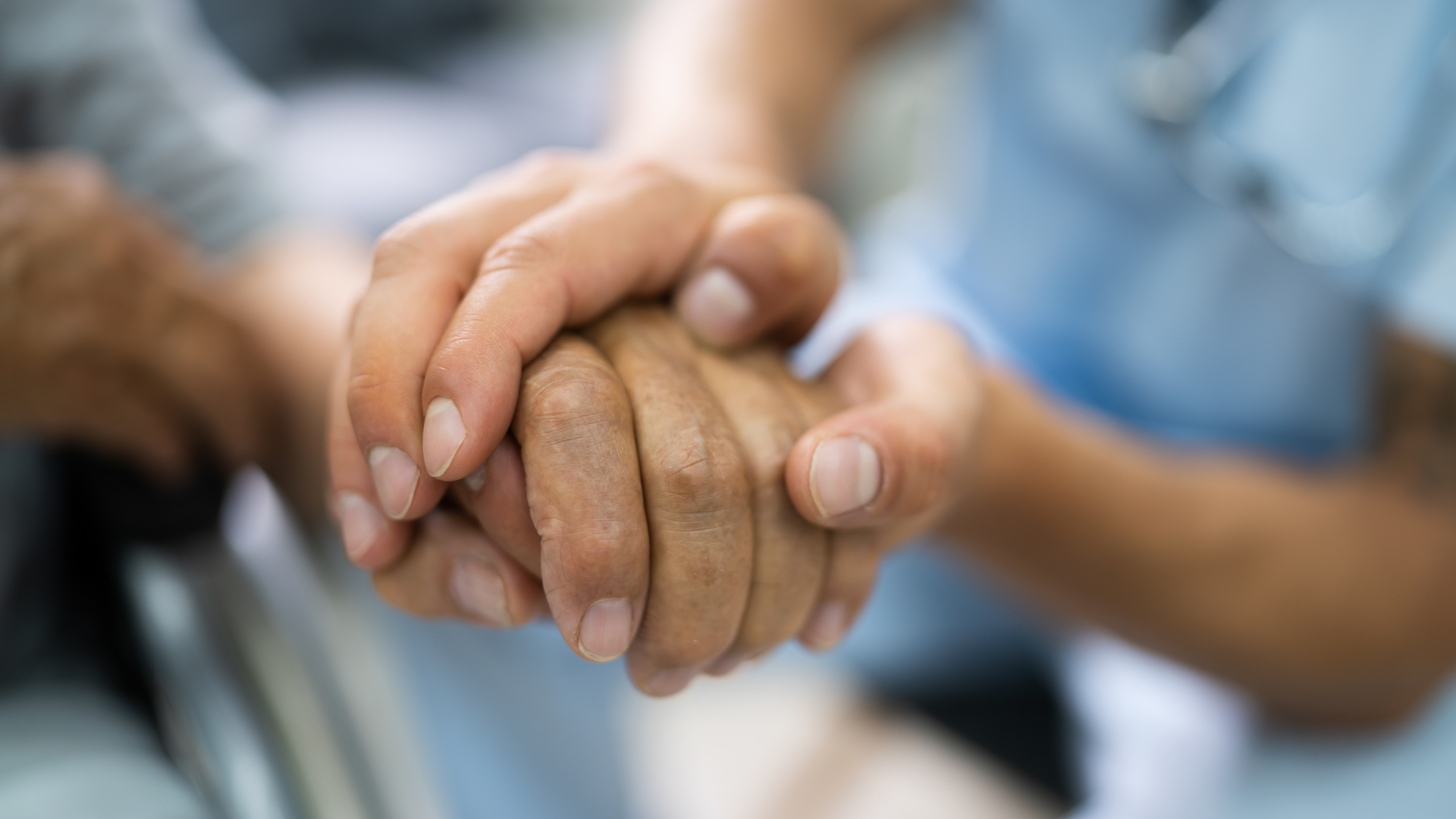 Person's hands held by another, possibly in a caregiving setting, offering comfort and support.
