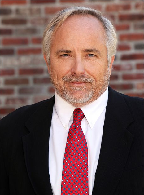 Man in suit and red tie, smiling in front of a brick wall.