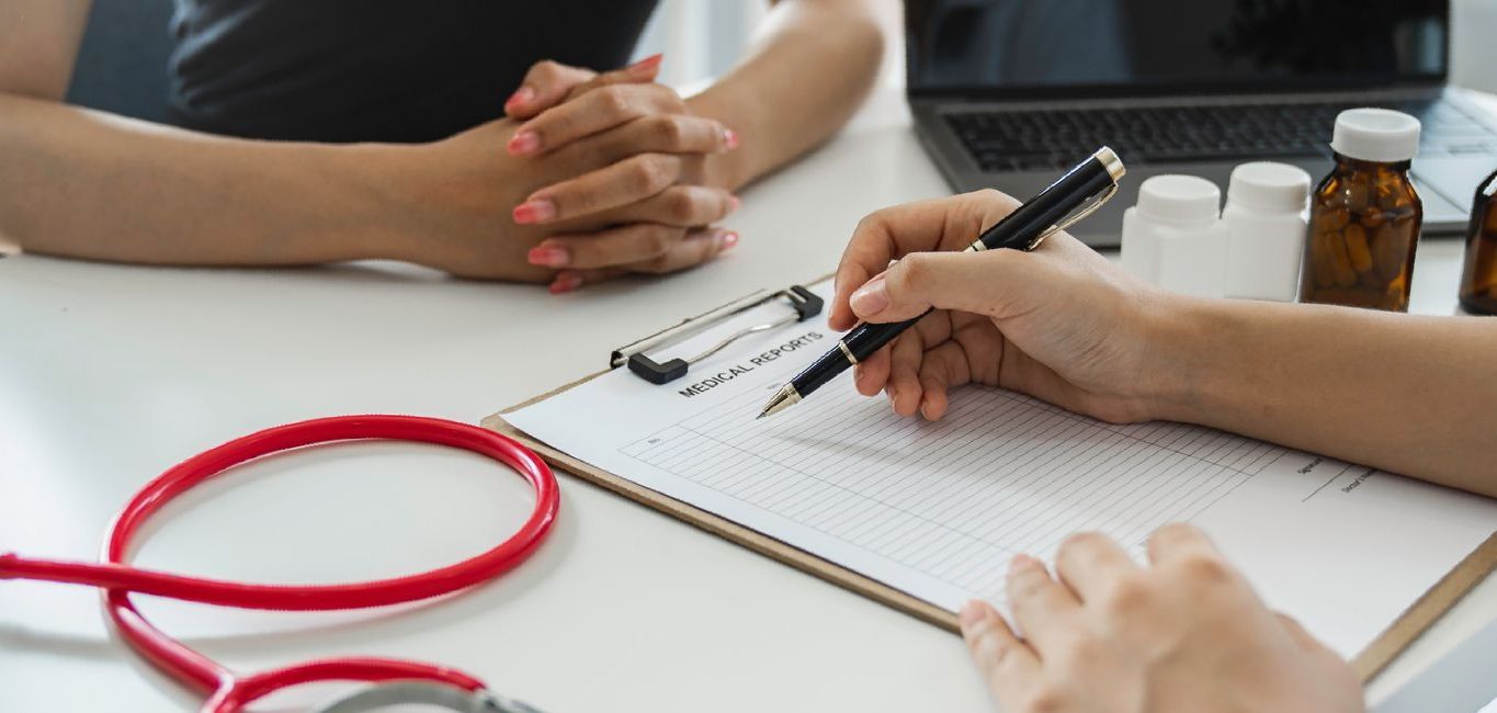 A doctor is sitting at a table with a patient and writing on a clipboard.