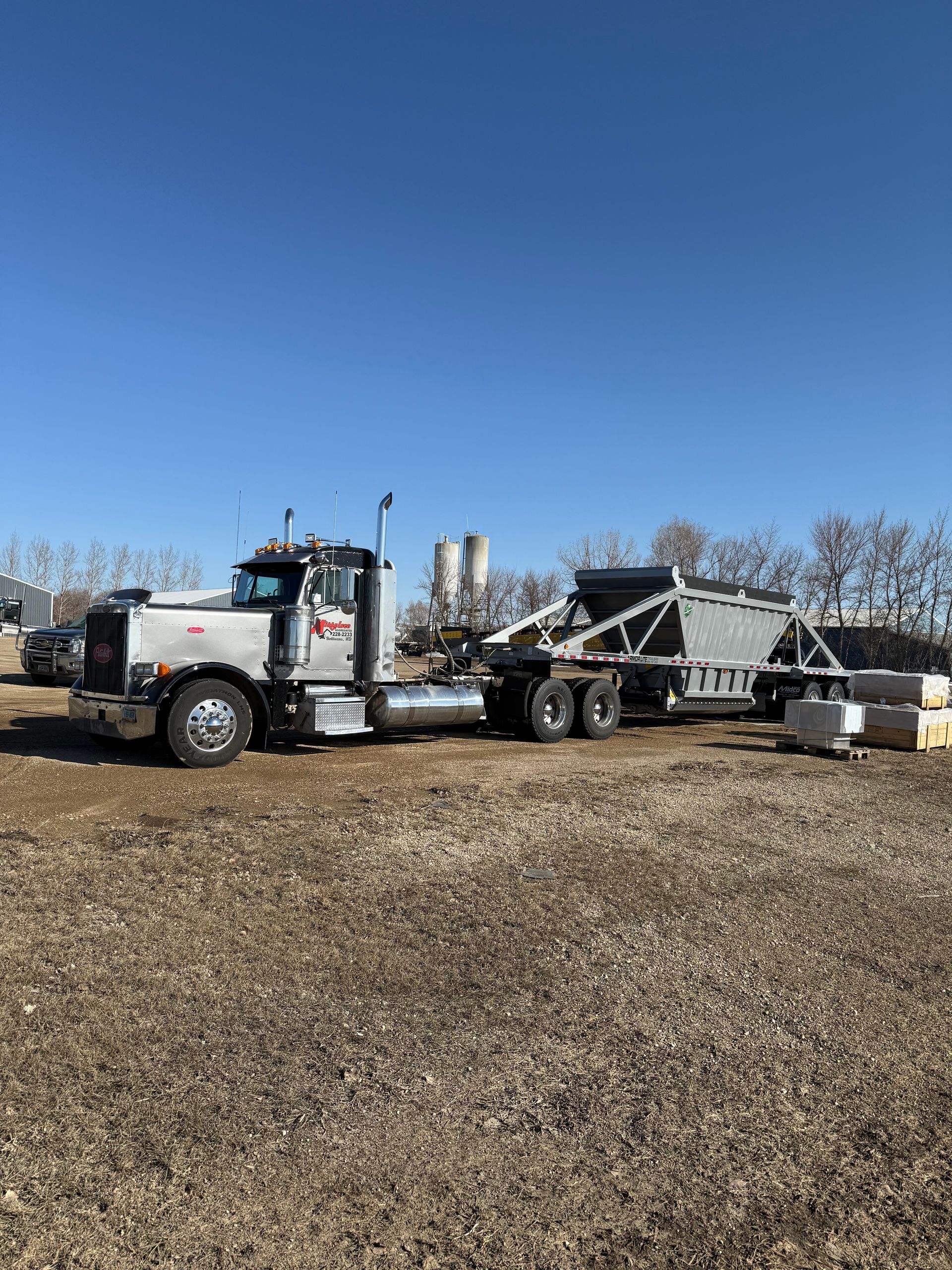 Semi-truck with trailer, gray and silver, parked in a rural area on a clear, sunny day.