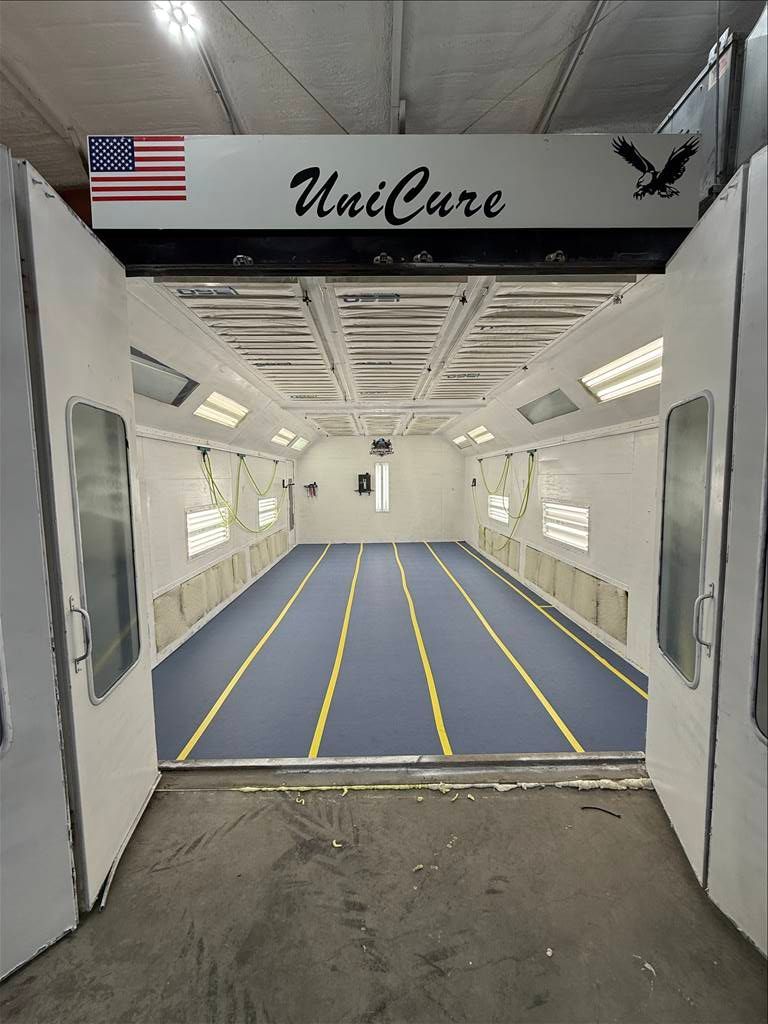 Inside of a UniCure spray booth with a blue floor and overhead lights. American flag and eagle logo above entrance.