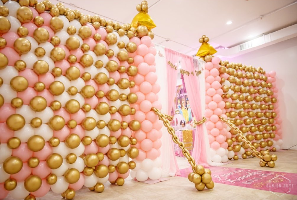 A wall made of pink and gold balloons with a castle in the background.