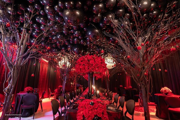 A room with tables and chairs and red flowers and black balloons on the ceiling.