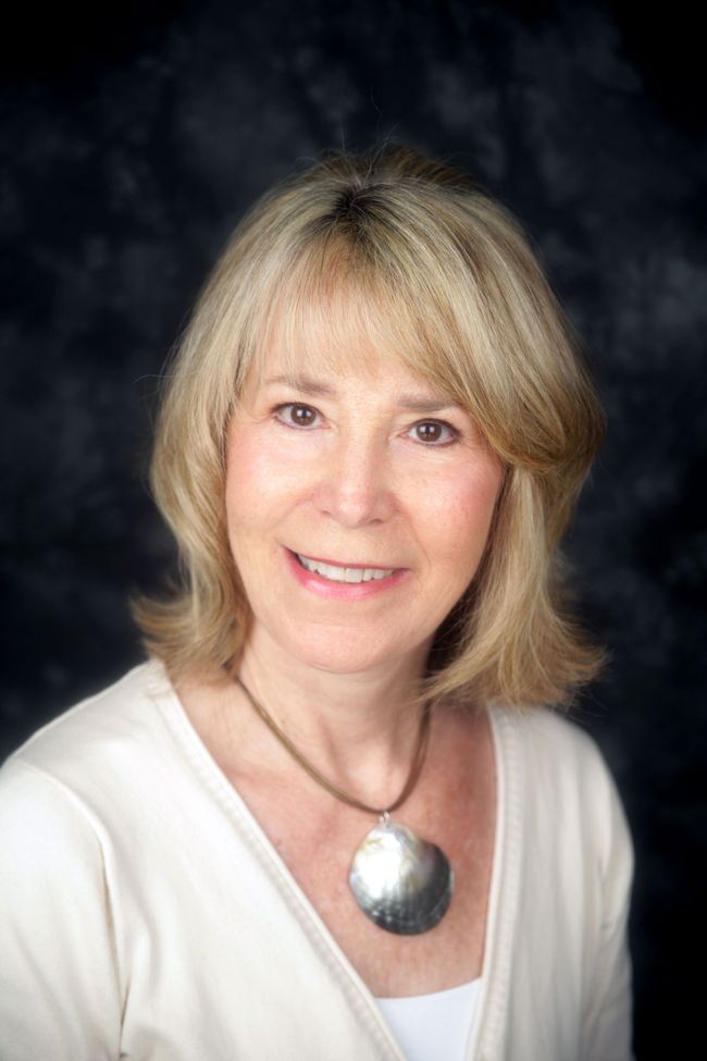 A person with shoulder-length blonde hair wearing a white top and a large, round metallic pendant necklace.