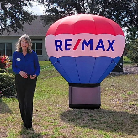 A woman stands in a yard next to an inflatable RE/MAX hot air balloon and a red open house flag.