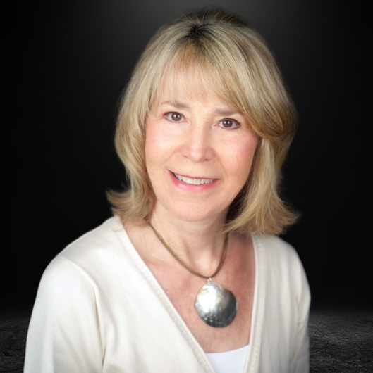 A person with shoulder-length blonde hair wearing a white top and a large, round metallic pendant necklace.