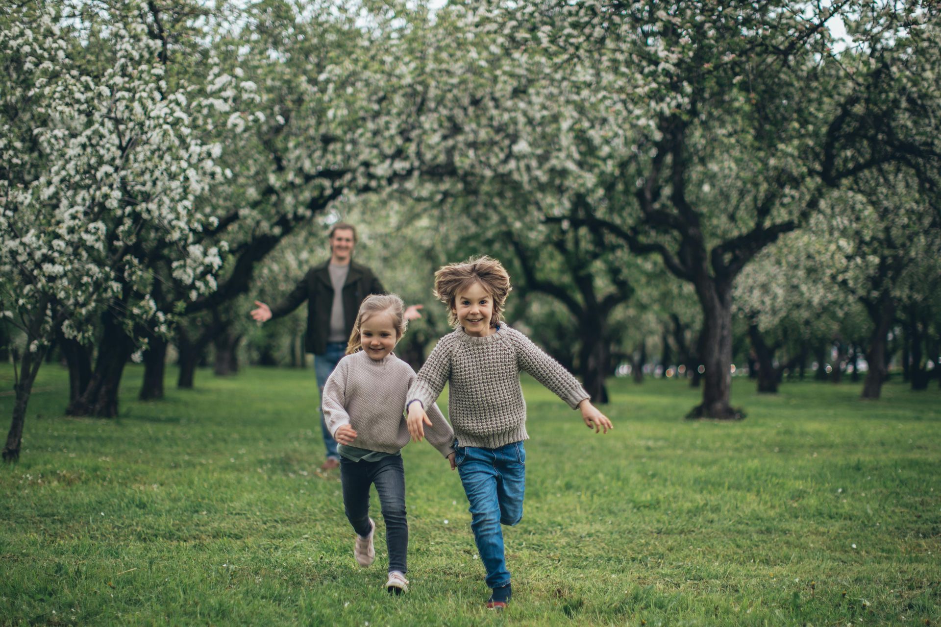 Two children run towards the camera through a blossoming orchard, a person smiles behind them.