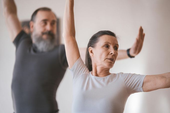 Two people in athletic wear practice a yoga pose with arms outstretched in a bright, indoor studio.