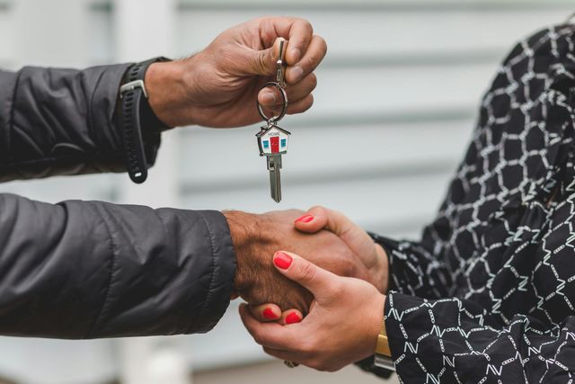 A person hands a house-shaped key to another person, symbolizing a real estate transaction.