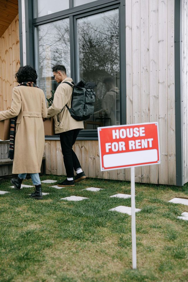 Two people carrying boxes walk toward a wooden house next to a red 