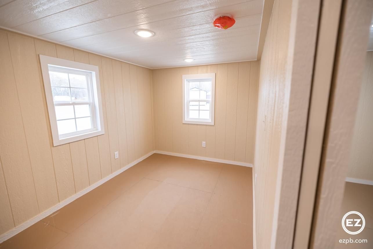 An empty room with two windows and a fire alarm on the ceiling.