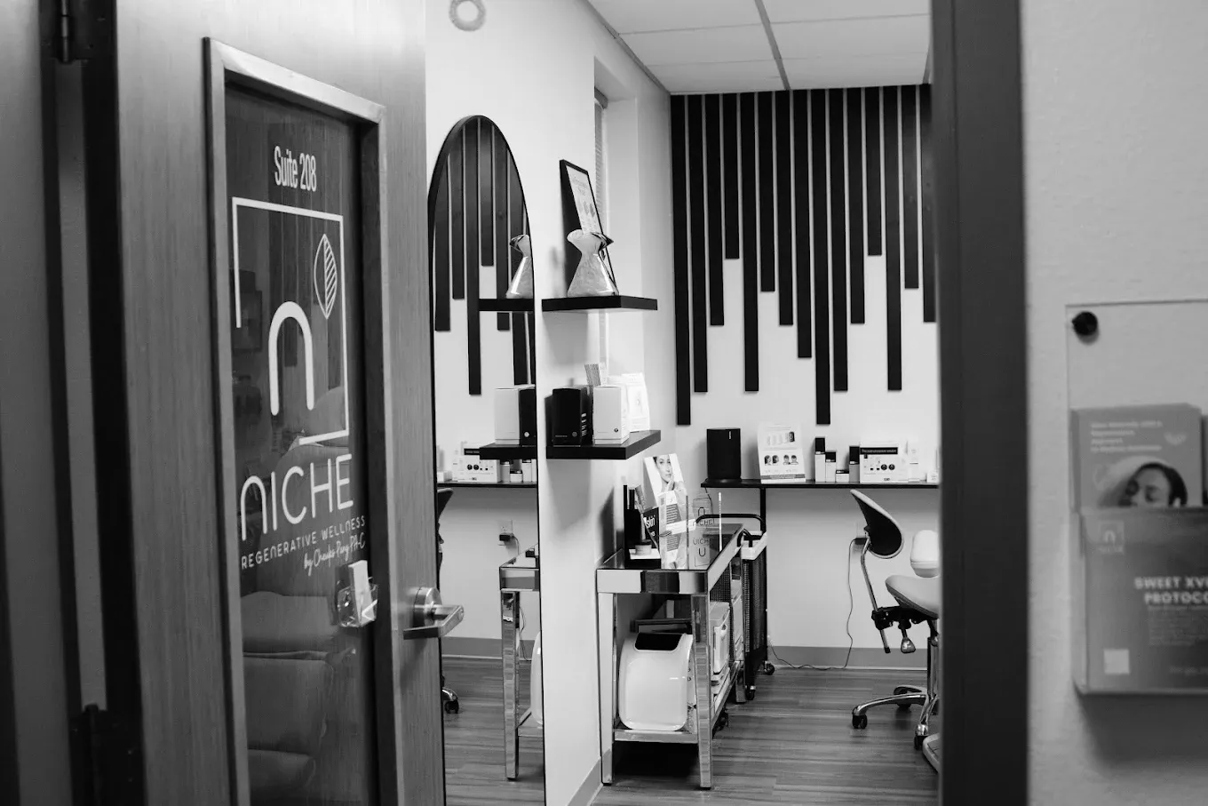 Interior of a salon suite with wood floors, black and white wall art, a styling mirror, and a desk, seen through a door.