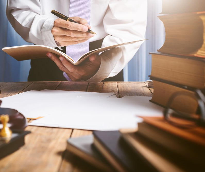 Person in business attire writing in a notebook on a wooden desk with books and paper. Sunlight shines on the scene.