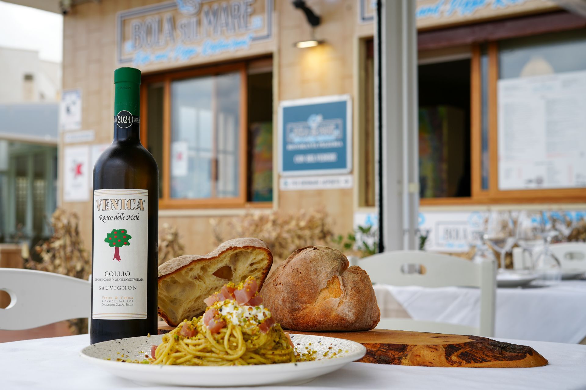 Tavolo apparecchiato per un pasto all'aperto in un ristorante con pasta, pane, bottiglia di vino e tovaglia bianca.