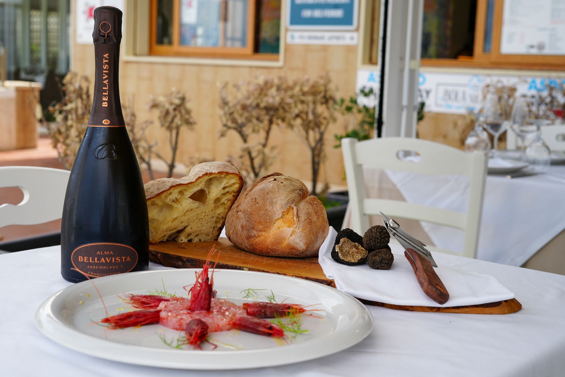 Tavola apparecchiata con spumante, pane, gamberi crudi, tartufi e interni di un ristorante.