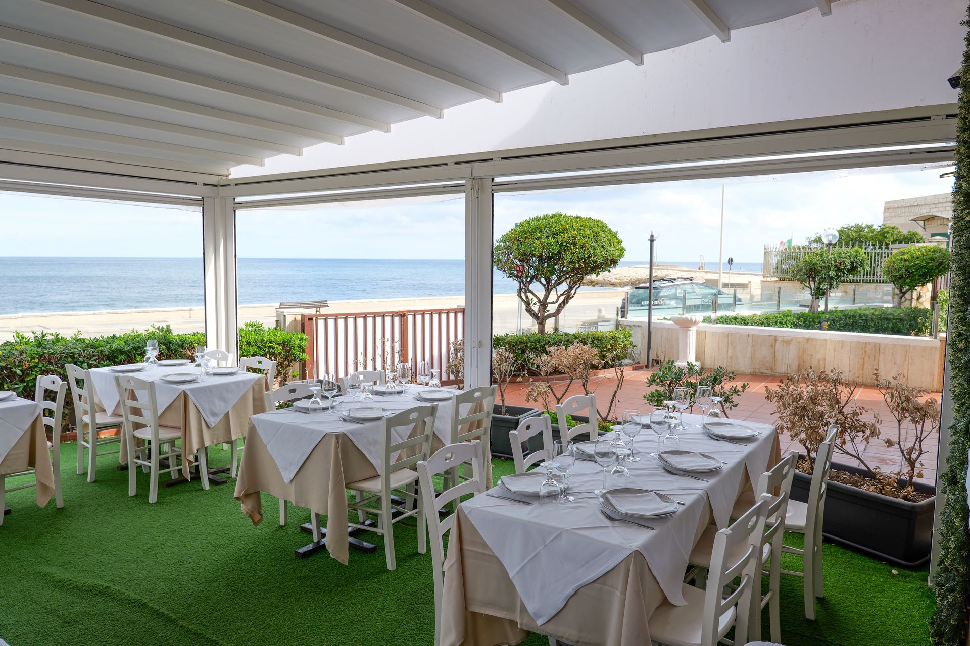 Patio esterno del ristorante con vista sull'oceano, tavoli apparecchiati, arredamento bianco e beige.