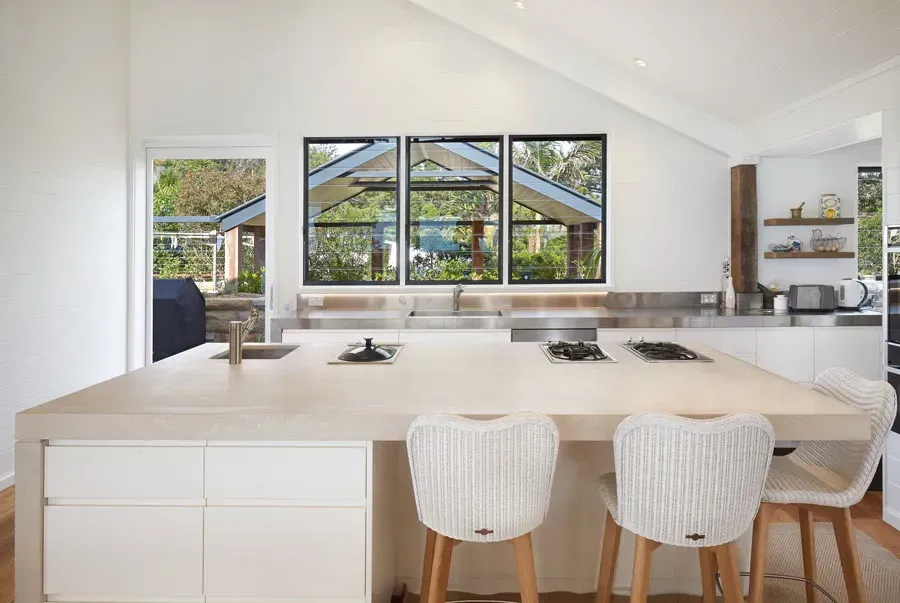 Bright white modern kitchen with island, two woven stools, and large windows overlooking greenery — Fineworx Joinery in Central Coast, NSW