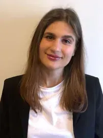 Woman with brown hair smiles, wearing a black blazer and white shirt.