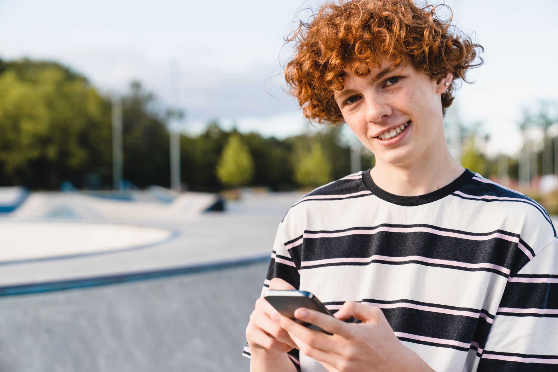Teenager with curly red hair and braces holding a phone, smiling at skate park.