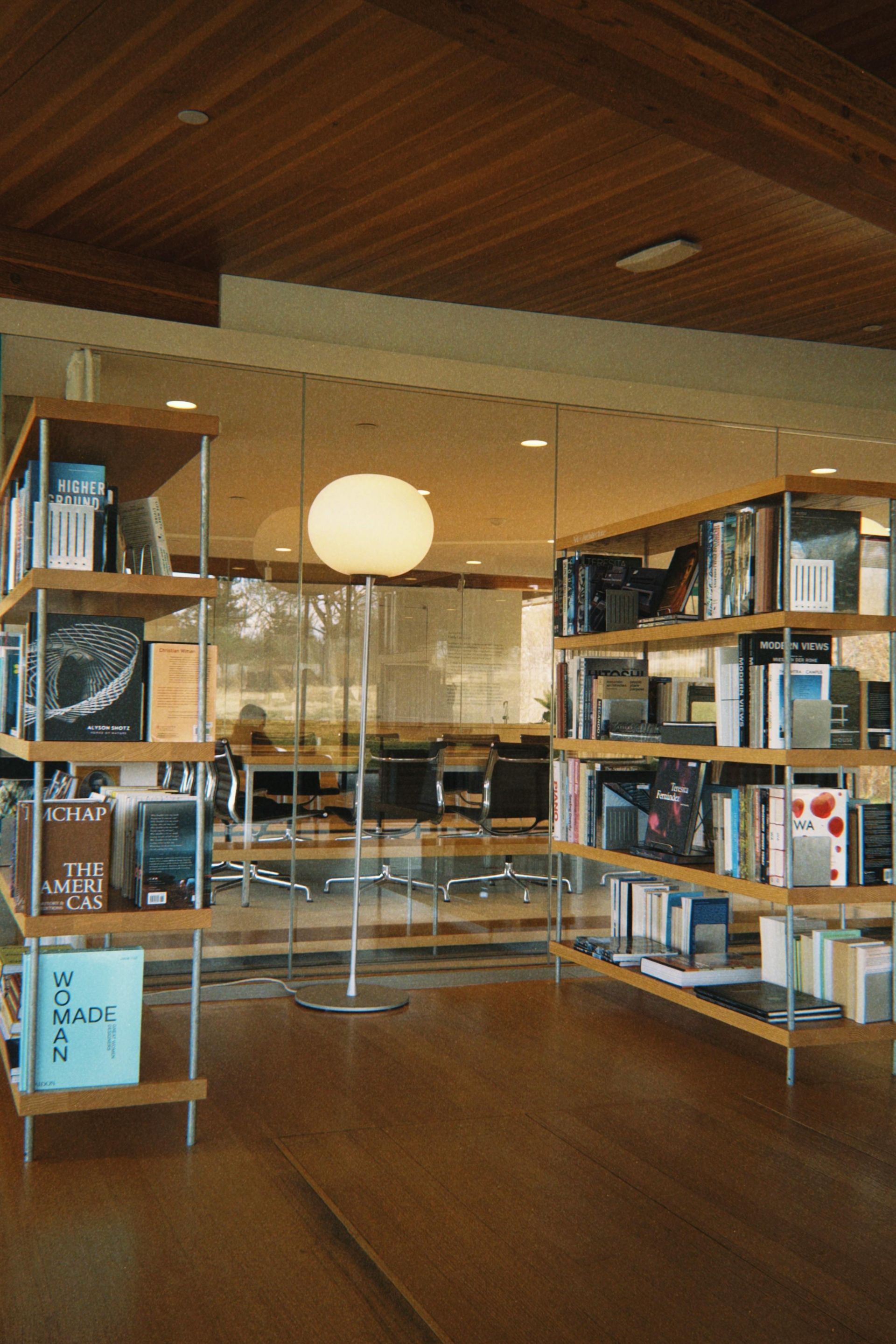 Book shelves with books in front of a glass wall and a floor lamp in an office.