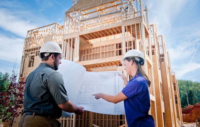 Two people stand looking at construction plans in front of a frame house.