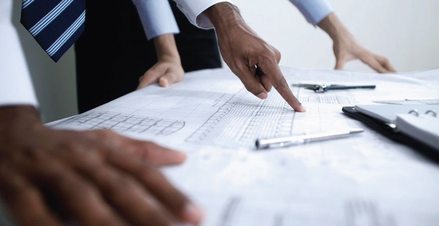 Some men look at plans on a table. One man points to something. 