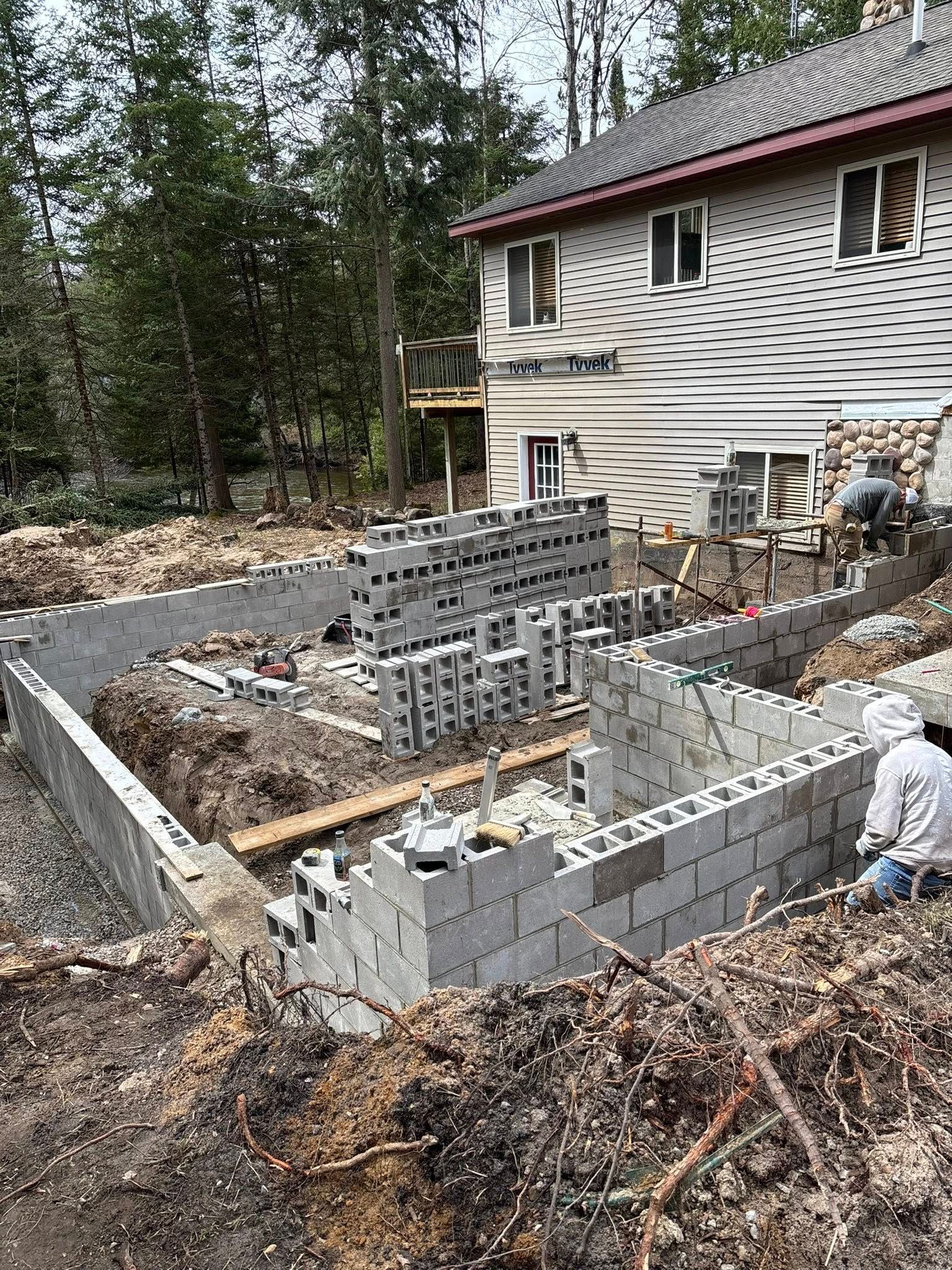 Construction of a concrete block foundation beside a two-story house. Workers are visible, surrounded by dirt and building materials.