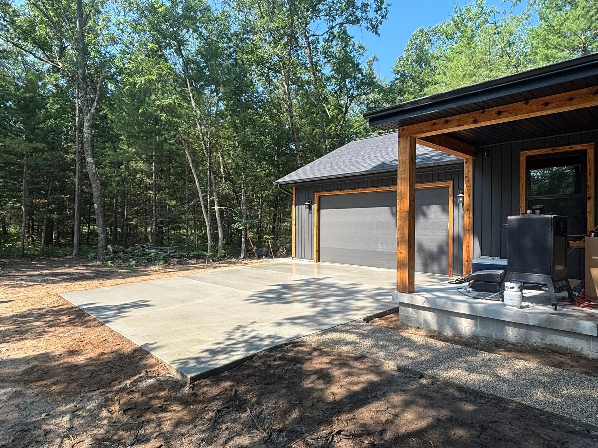 A newly constructed gray garage with a concrete driveway in a wooded setting. The building has dark gray siding and a wooden porch.