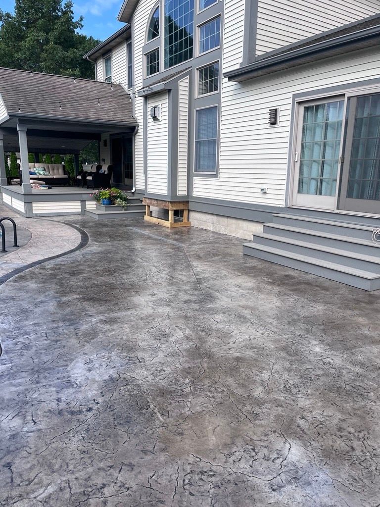 A newly poured concrete slab in an outdoor setting, surrounded by cinder blocks and dirt, with a house and shed in the background.