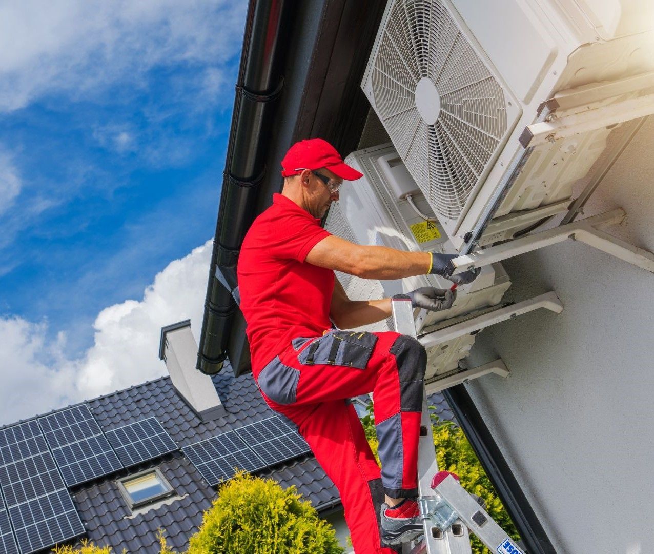 A man is standing on a ladder fixing an air conditioner on the side of a building.