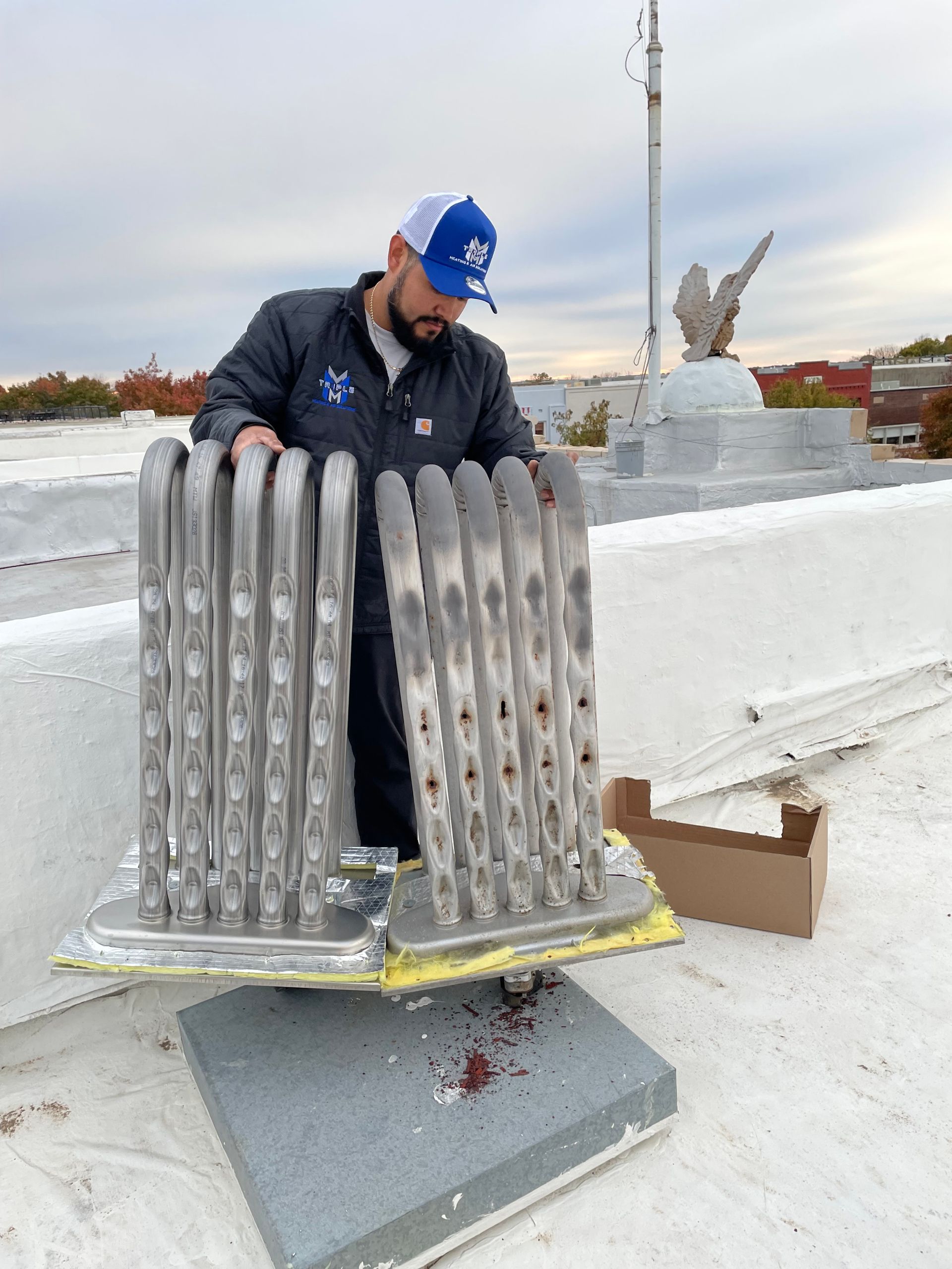 Man on rooftop, working on equipment. Gray, tubular structure. Blue hat, jacket. Overcast sky.