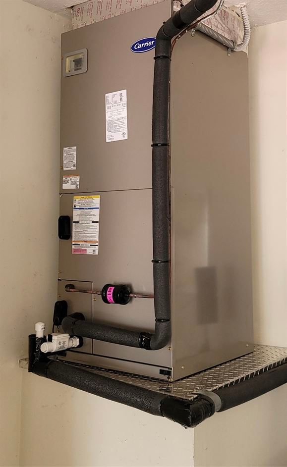 HVAC unit in a corner, with insulated pipes. The unit is gray and sits on a metal shelf.