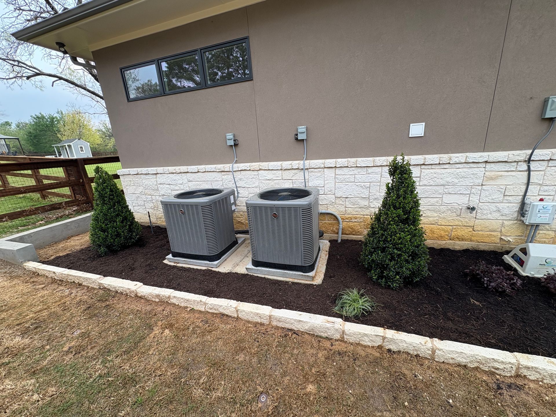 Two gray air conditioning units next to a tan building with landscaping and trees.