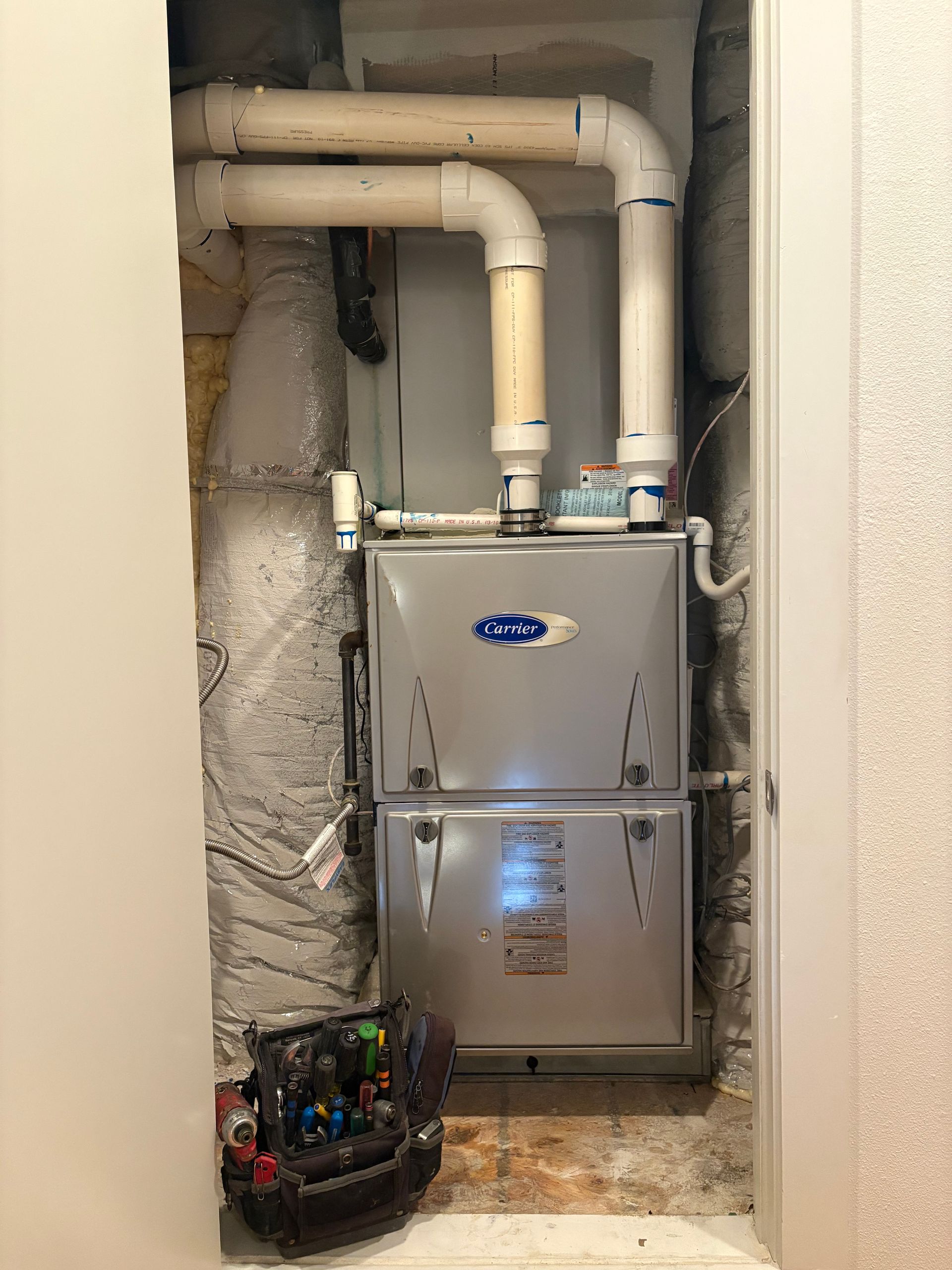 Furnace in a closet, with white vents and a tool bag on the floor.