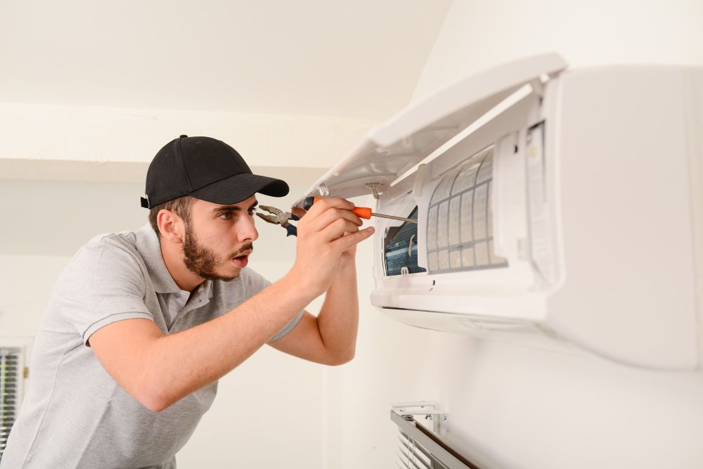 HVAC technician in cap, inspecting and repairing a white air conditioning unit on a white wall.
