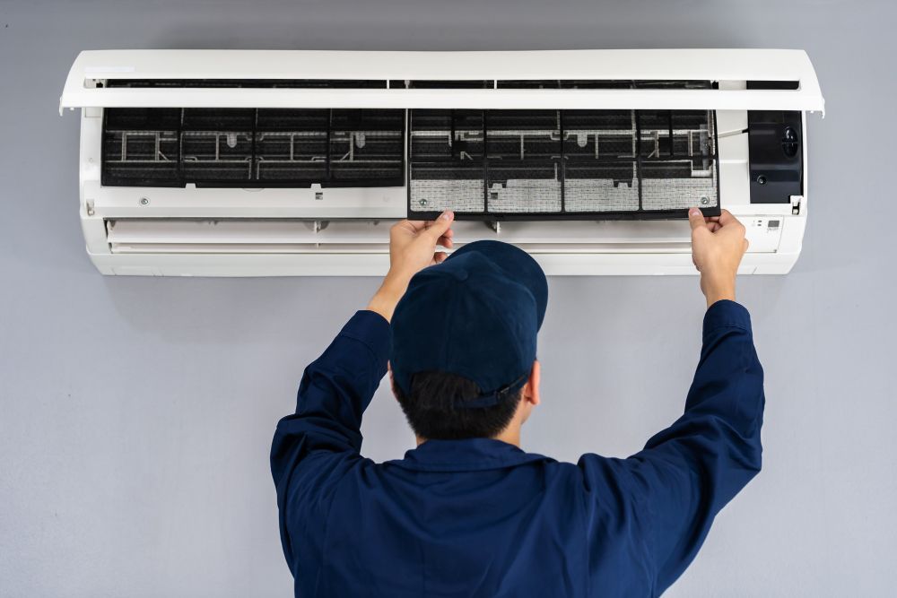 Person in blue uniform removing air filter from an AC unit mounted on a gray wall.