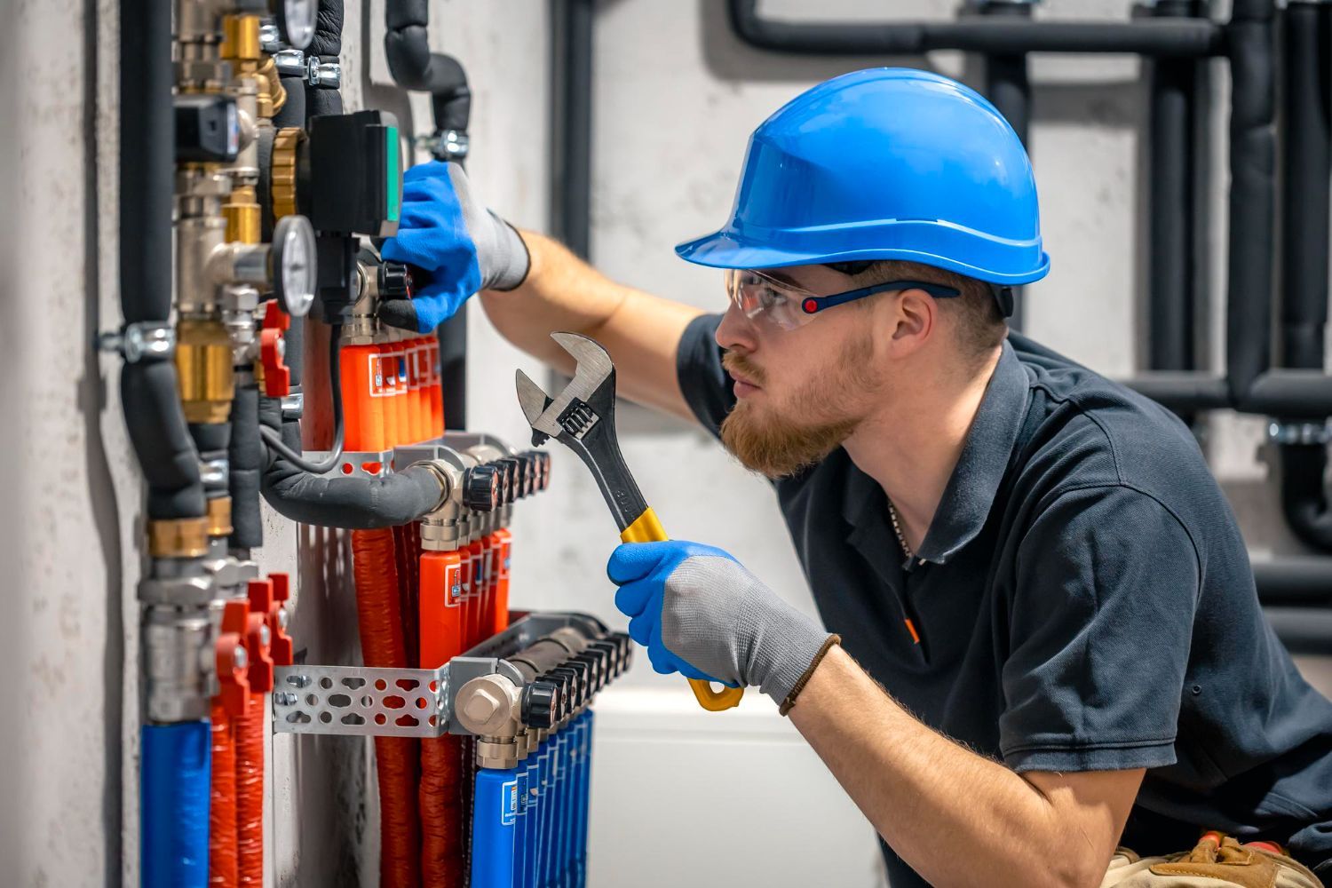 Plumber in a blue hard hat, gloves, and safety glasses working on water pipes with a wrench.