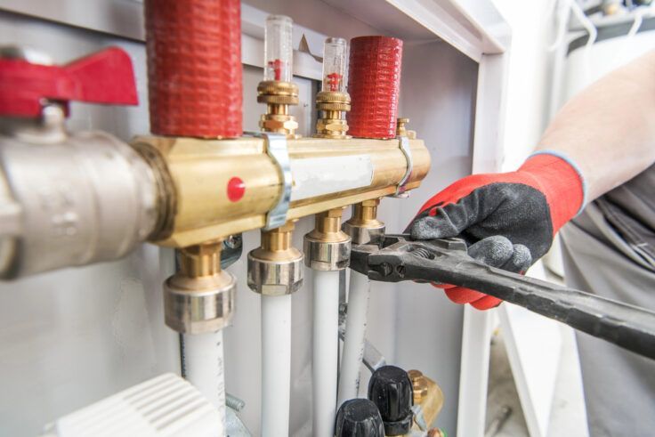 Plumber using a wrench on pipes. Red and black gloved hand in focus near brass fixture and white pipes.
