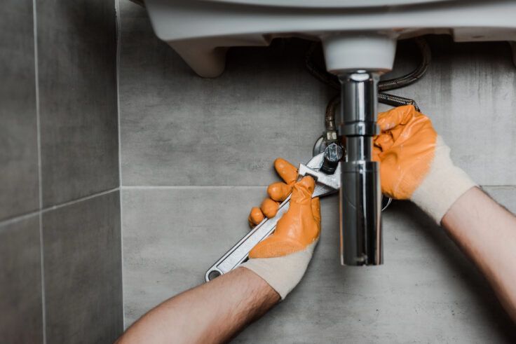 Hands with orange gloves using a wrench under a sink.