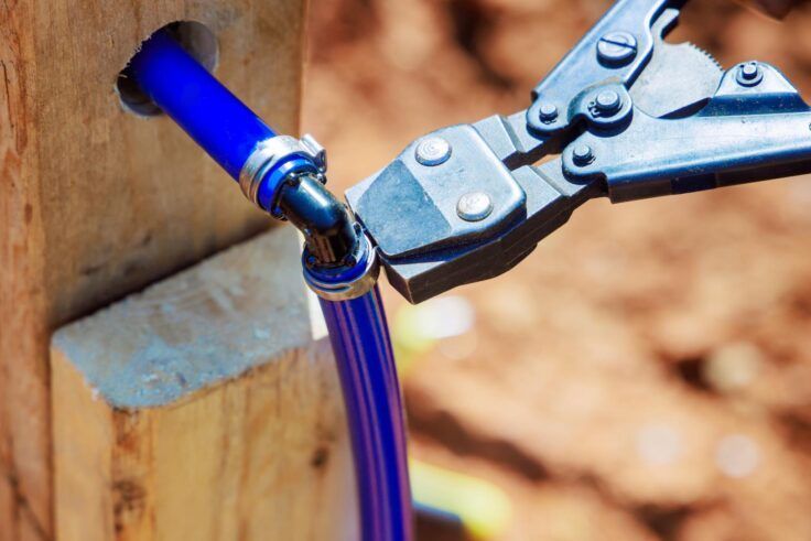 Bolt cutters cutting a blue pipe connected to a black fitting protruding from a wooden post.