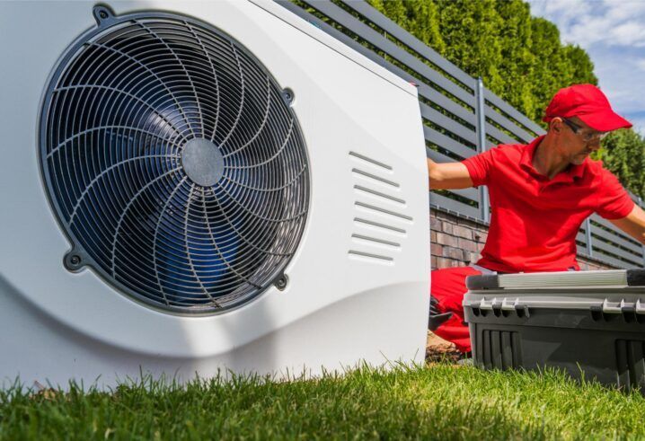 HVAC technician in red shirt and cap works on white heat pump unit, green grass, gray tool box.