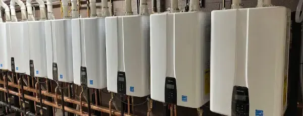 Row of white tankless water heaters with black control panels, copper pipes, and a grey background.